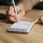 From above of crop faceless male student writing with pen in notebook at wooden desk during lesson